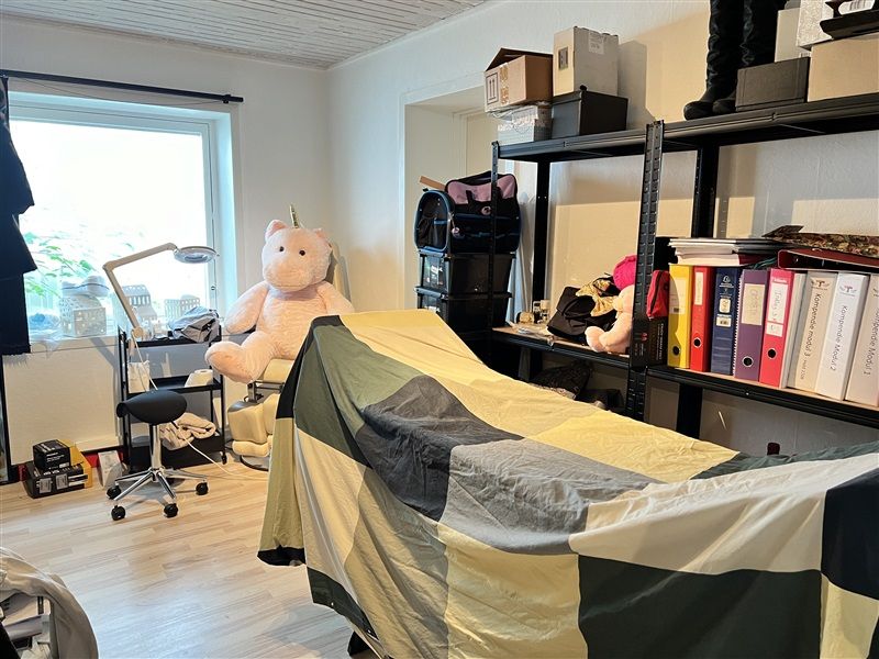Et værelse med et tæppe, hvor en teddybjørn sidder, samt flere bøger og dokumenter på et højt hylde