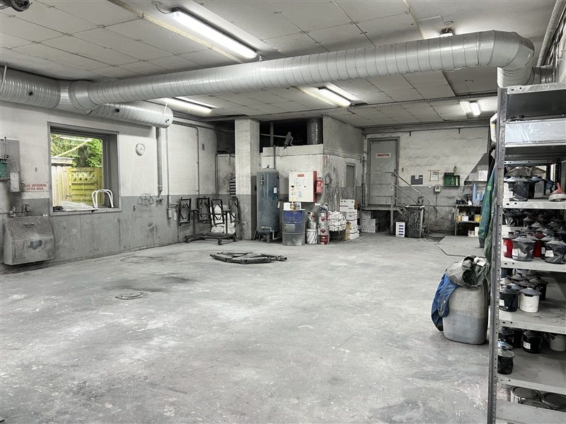 Industrilokaler med to store vinduer, lysekroner og ventilationsrør på loftet og tomme stel samt udstyr og malingskanister