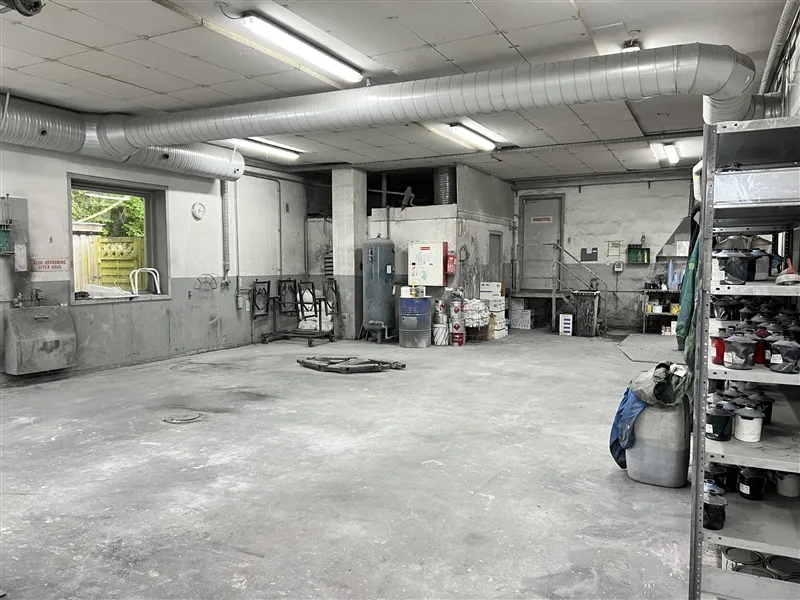 Industrilokaler med to store vinduer, lysekroner og ventilationsrør på loftet og tomme stel samt udstyr og malingskanister