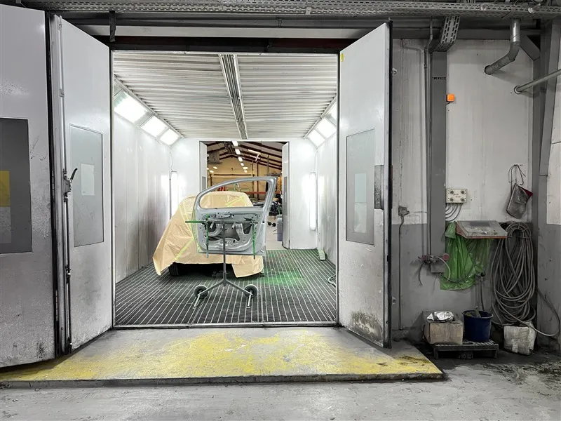 Erhvervslokaler med åbne døre og bilramme på hjulstativ i spraykabine med hvide vægge og metalhylder