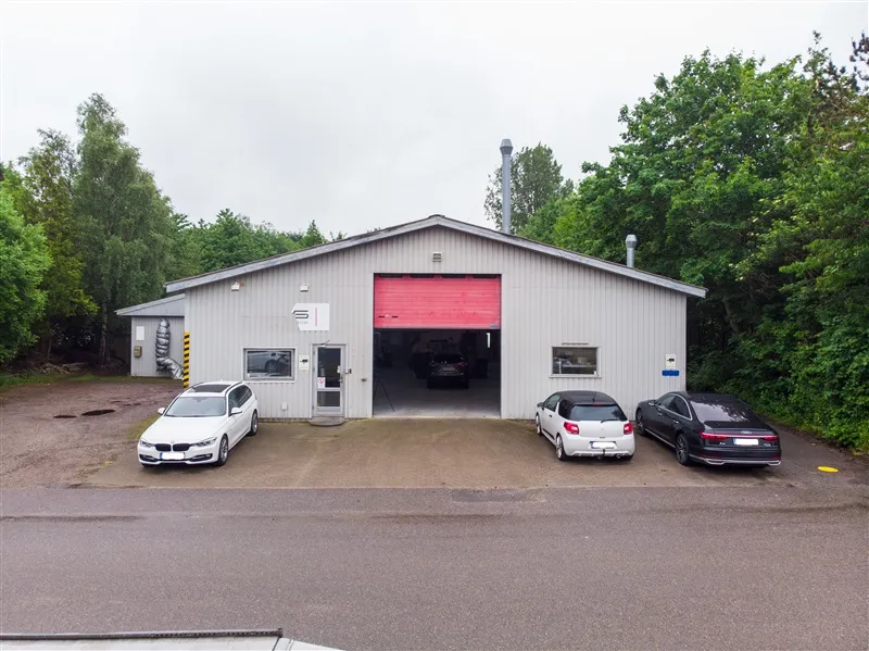 Foran en bygning med garagedører og biler er der tre parkerede biler og et træskovet baggrund