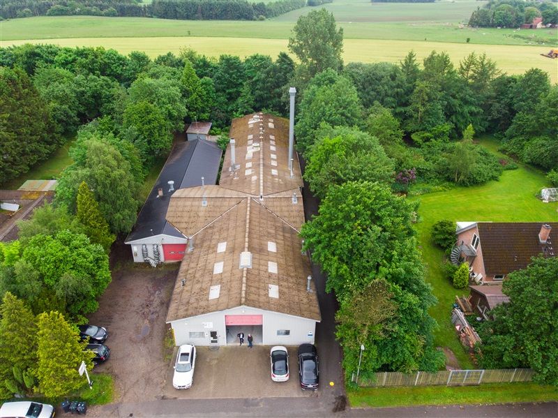 Høj bygning med stort tag og garagedør står på en grund med parkerede biler og grønne træer og buske