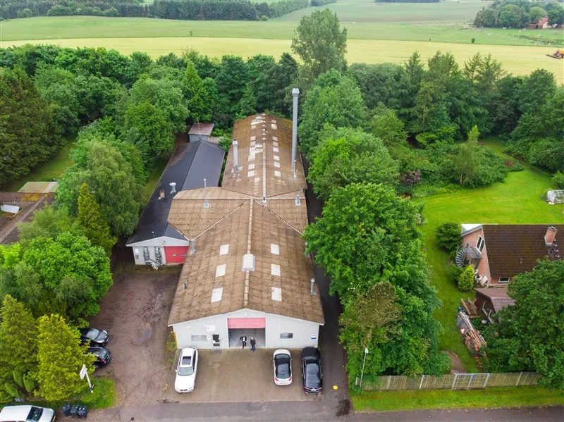 Høj bygning med stort tag og garagedør står på en grund med parkerede biler og grønne træer og buske