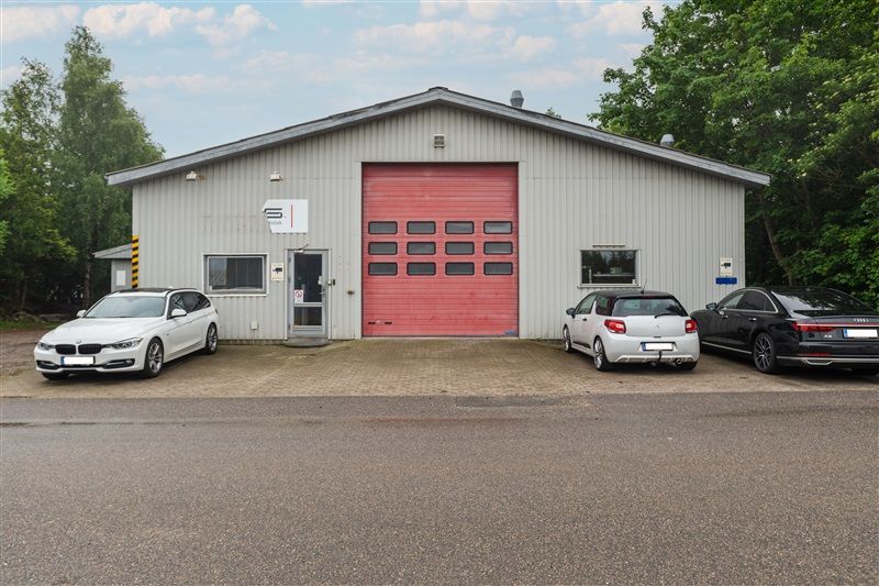 Erhvervsejendom med rødt garagedør og tre parkerede biler på forsiden og tre træer i baggrunden