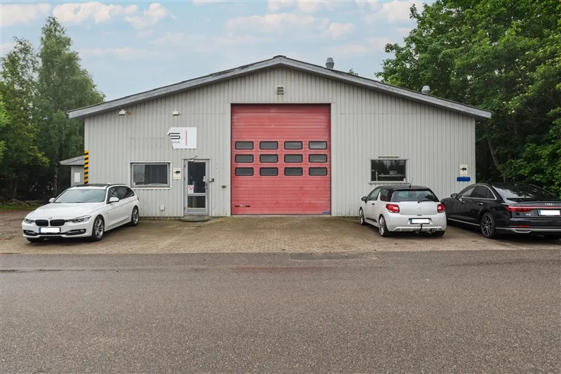 Erhvervsejendom med rødt garagedør og tre parkerede biler på forsiden og tre træer i baggrunden