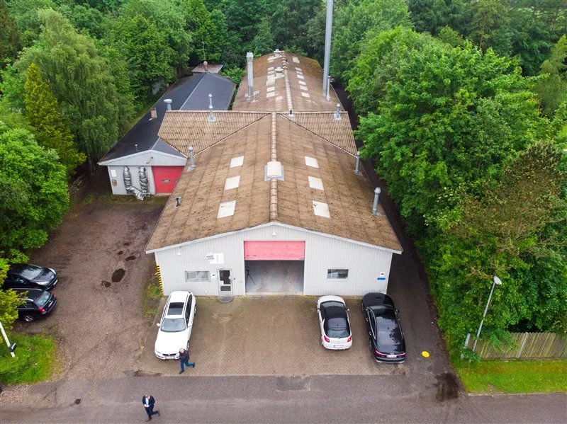 Personer i forgrunden går mod et hvidt hus med garage. Biler parkeret foran hoveddøren. Grønne træer og en lysstang i