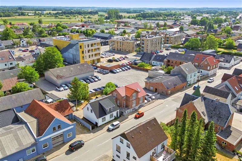 Gade med bygninger og parkeringsplads, biler parkeret, en grøn buske og træer