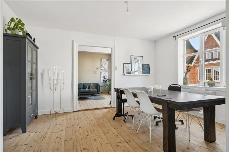 Moderne stue med massivt trægulv og hvide vægge, massivt træbord og hvide stole og lysekroner, plante og fotorammer på væggen