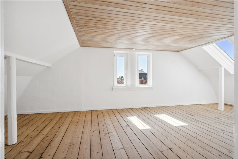 Foto af en rummelig loftsværelse med lyse trægulve og hvide vægge, der reflekterer lys fra vinduerne