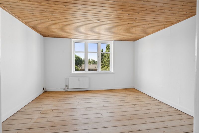 Foto af et tomt og lyserum med hvid maling på væggene og trægulv og et vindue med udsigt til grønne træer