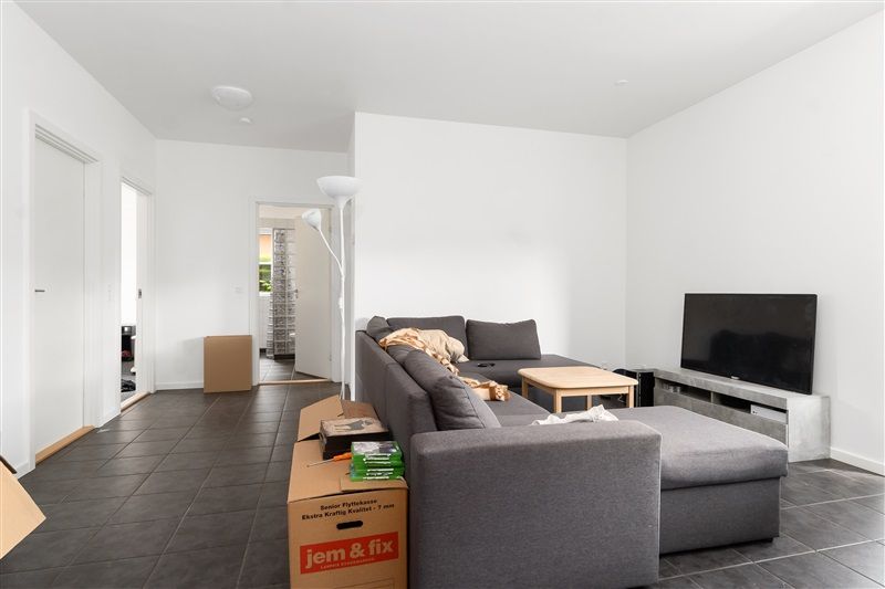 En stor fjernsynsskærm står på en høj hylde, og der er en sofa med et bord og nogle kasser i stuen