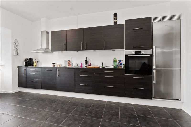 Moderne køkken med mørke skabe, hvide vægge, rustfri køleskab og indbygget ovn samt en række køkkenredskaber og krydderier