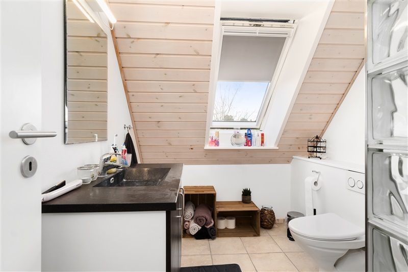 Foto af et badeværelse med hvide vægge og loft. Der er en toilet, et vindue og et spejl med lyse og trævægge.
