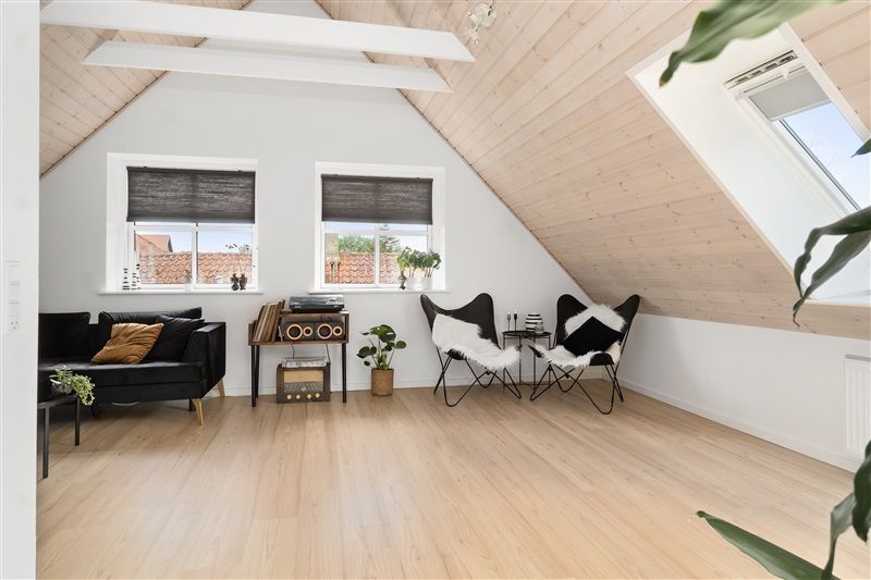 Et luksuriøst loftsværelse med højt loft, lyse gulvbelægning, og moderne møbler inklusiv et sort sofa udstyret med puder og