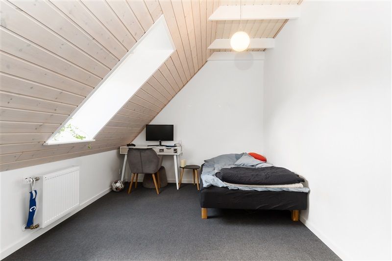 Et lille soveværelse med enkeltseng, skammel, skrivebord og computer samt en hvid radiator på væggen