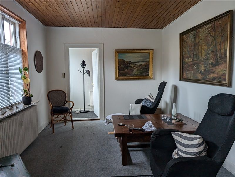 Billede af et lyse og moderne stue med træloft, hvid væg og trægulv. Et bord med en flaske og en vase og to møbler med