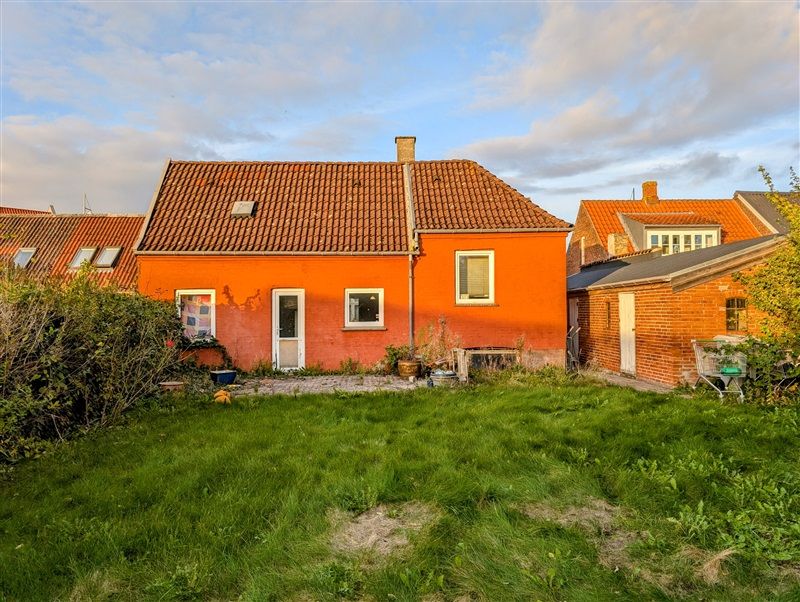 En gul tegltagget bygning med plantepotter udenfor og en grøn græsplæne i forgrunden