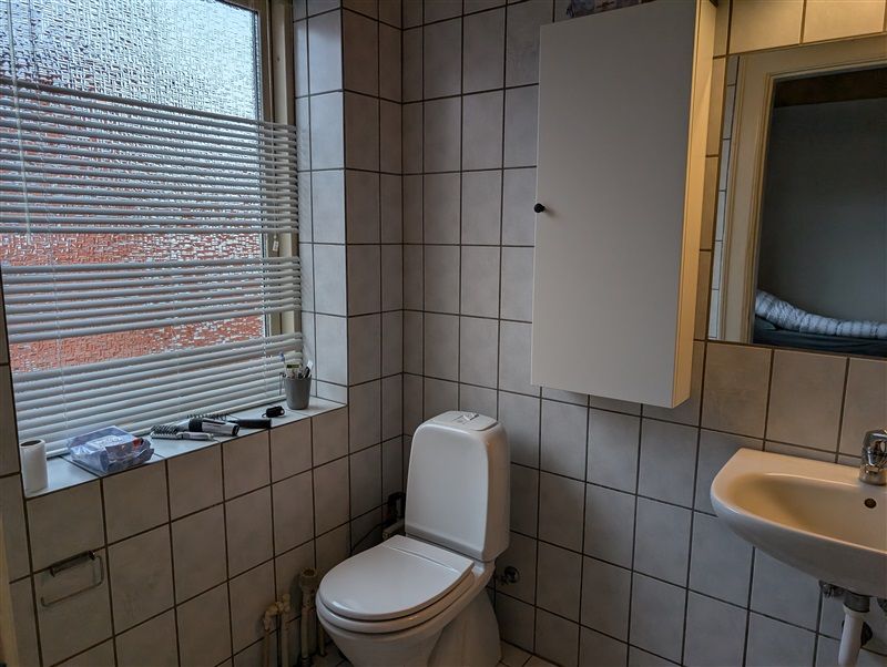 En lille badeværelse med toilet, håndvask, spejl og vindue. Toilettet er placeret ved siden af håndvasken og spejlet.