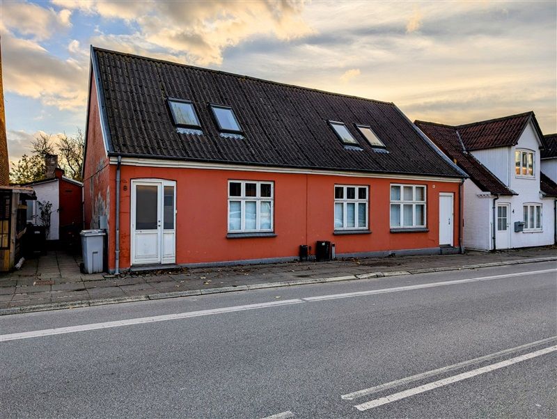 En rødmalet bygning med sorte tag og lyseruder er placeret på hjørnet af en tomgangsvej i Middelfart