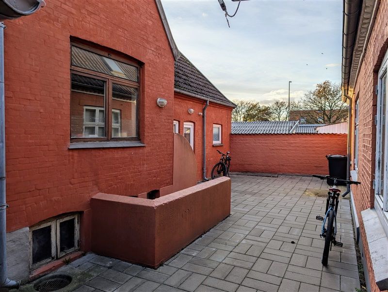 To røde bygninger med vindue og dør og to cykler parkeret i baghaven med stengulve