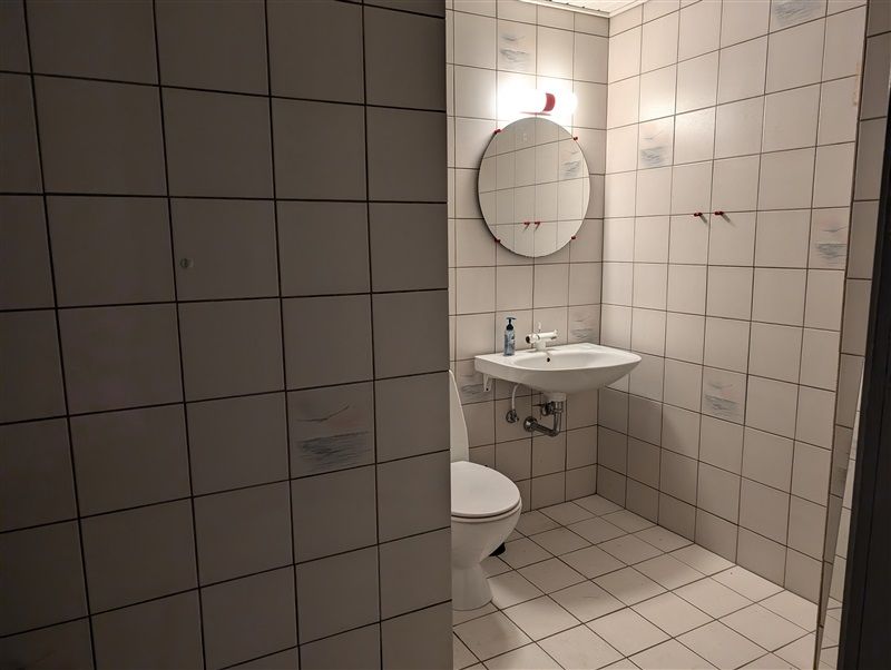 Et lille badeværelse med hvidt flisede vægge og gulv samt et toilet og en hvid håndvask med en spejl