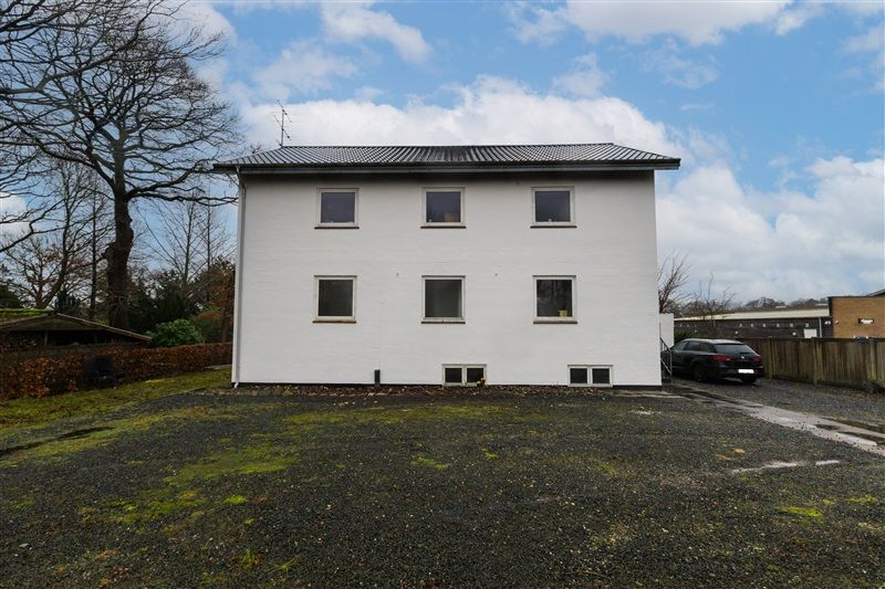 En stor hvid bygning med flere vinduer og en parkeringsplads foran er parkeret en sort bil