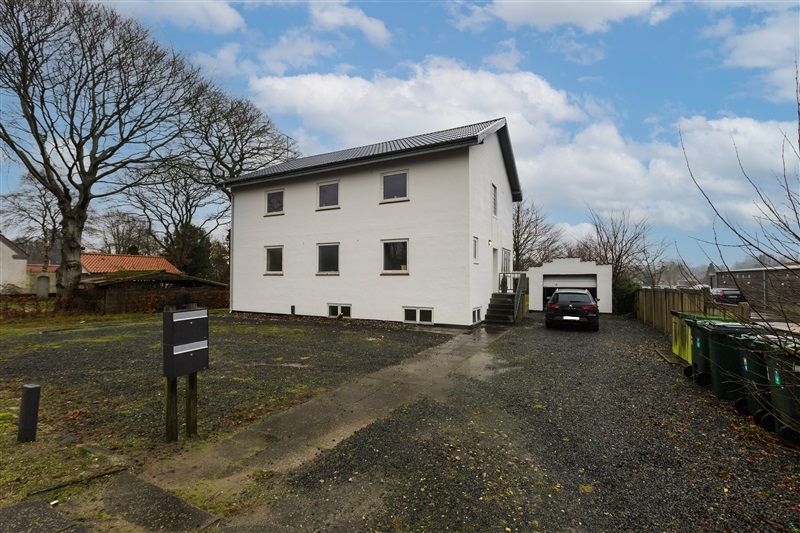 Et hvidt hus med garage og trapper udenfor, i en forstadsområde med grønne træer og parkerede biler