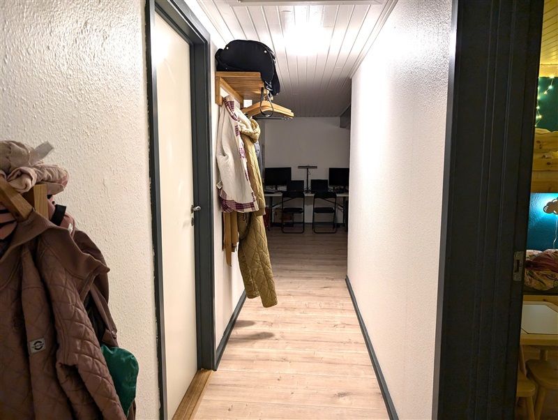 En gange med to stole, computere og en hængehylde med en sort taske og tøj.