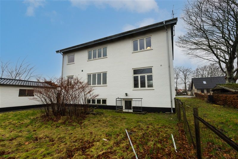 En hvid to-etagers bygning med flere vinduer, en indgangsdør og et lille græsområde med en skur og en stærk tagrenne