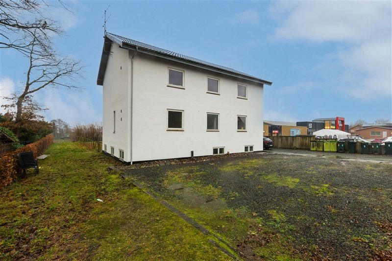 En hvid bygning med fire vinduer og en række skraldcontainere på grunden