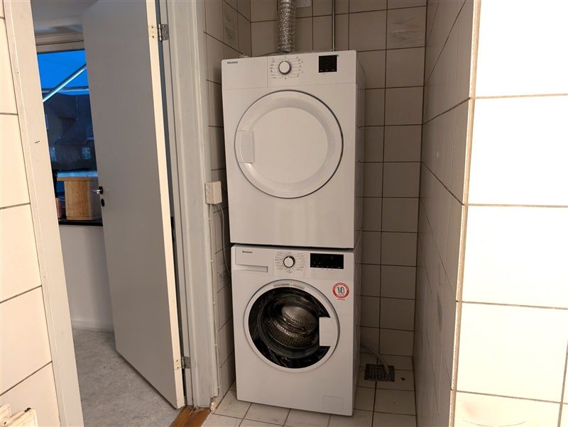 To hvide tøjvaskere står sammen i et lille badeværelse med hvide kakler og åben dør