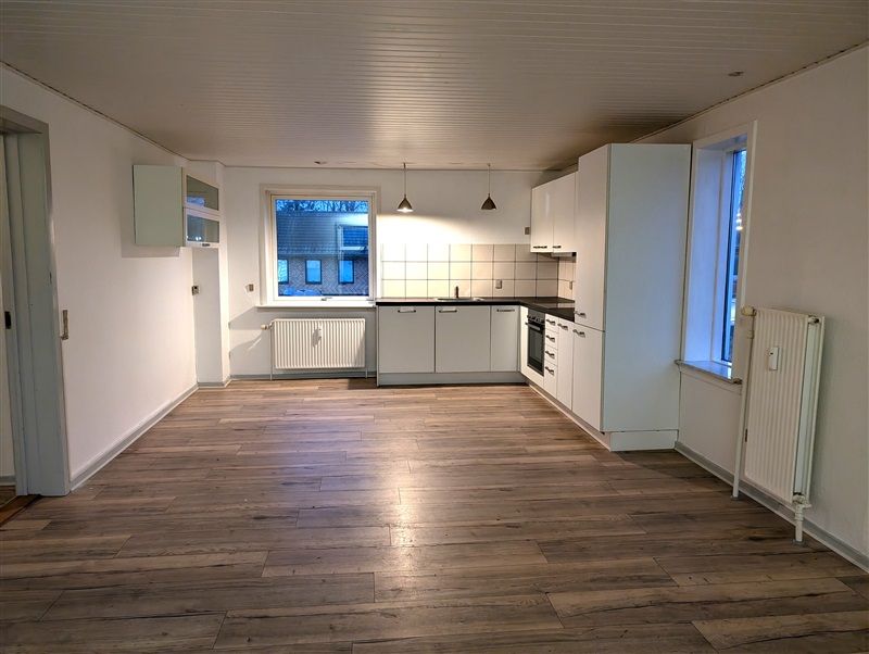 En tom lejlighed med moderne køkken og lyse lofter, hvor trægulve og hvide vægge skaber en hyggelig atmosfære