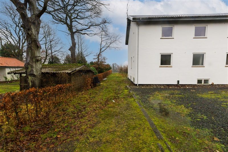 Et hvidt hus med flere vinduer står ved siden af en vej og en lille grønsnethæk og træer