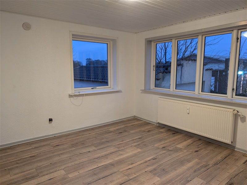 Et tomt rum med trægulve og store vinduer tilbyder udsigt til gårdsplads og et hus i baggrunden