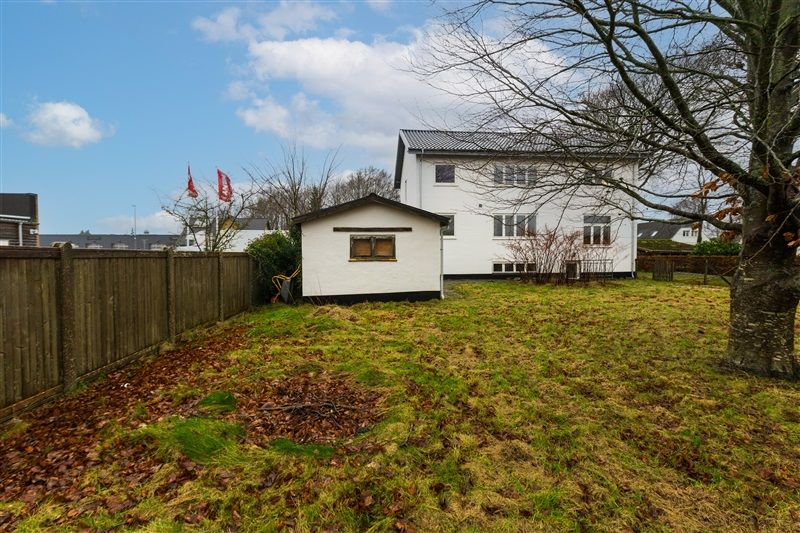 Et hvidt hus med rødt tag og røde faner står i en have med græsplæne og et lille sommerhus. En træstøtte med flagpole og en