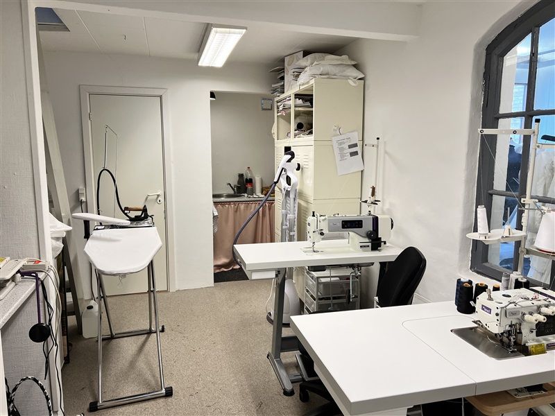 Et værksted med to symaskiner og en strygebræt, en strygejern samt flere kasser med forsyninger og et skab med tekstiler