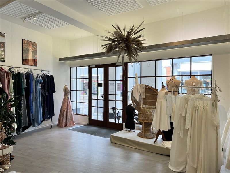 En butik med kjoler og tøj på strygebrætter og manequins samt lysekroner og plante i forgrunden