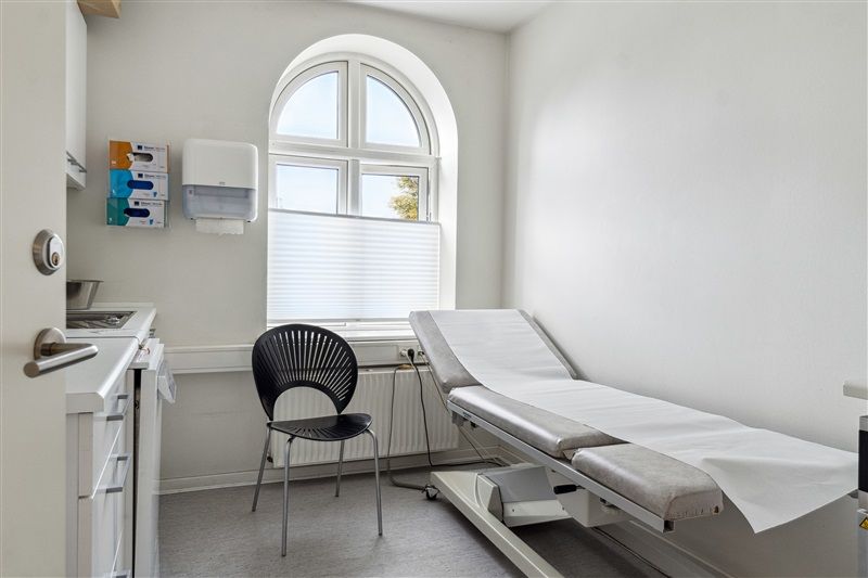 En tom lægepraksis med hvidt udstyk, runde vinduer, en hvid senge og en stol placeret nær en radiator