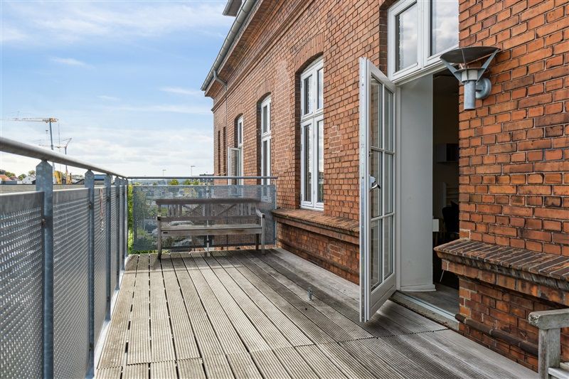 Terrasse med trægulv og lyserøde mursten. Balkon med metallynløb og åben dør. Søjle og lysekrone med udsigt over landsbyen.