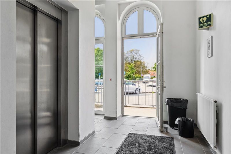 En indgangsdør til et lejlighedsbyggeri med elevator, 2 skraldespande og en hvid radiator