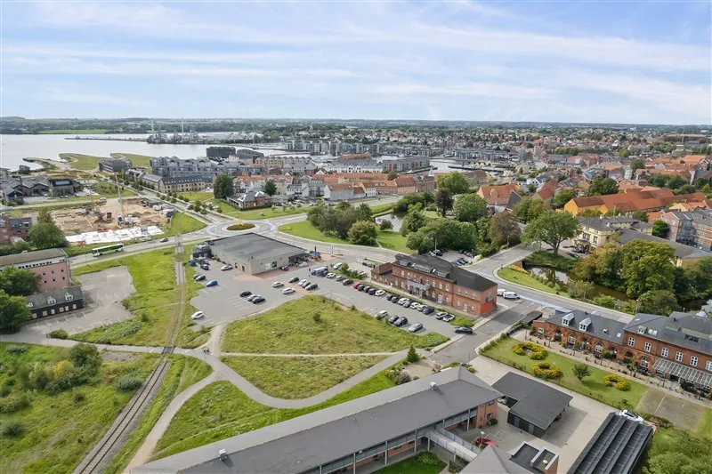 Billede af byen Nyborg med flere bygninger og parkeringspladser, tæt på vandet, med grønne træer og busser