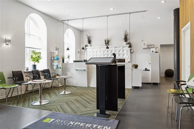 En moderne lobby med en lysegul væg og en gulvtæppe med teksten RycXperten. Her er en bord med et rødt bord og stole samt et