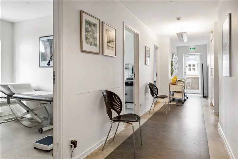 Kort korridor med hvide vægge og gulv og flere stole og møbler samt lægebord