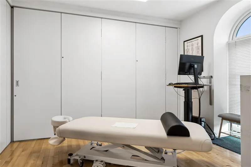 Behandlingsrum med massagebord, stol, computer og hvidt kabinet med to døre og en åben dør