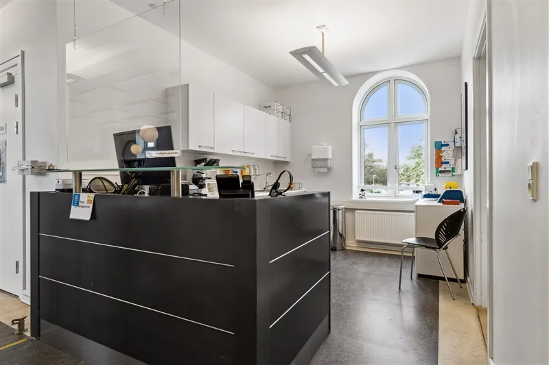 Kontor med høj glasvæg og sort skranken, skrivebord, computer og stol samt vinduer og radiatorer