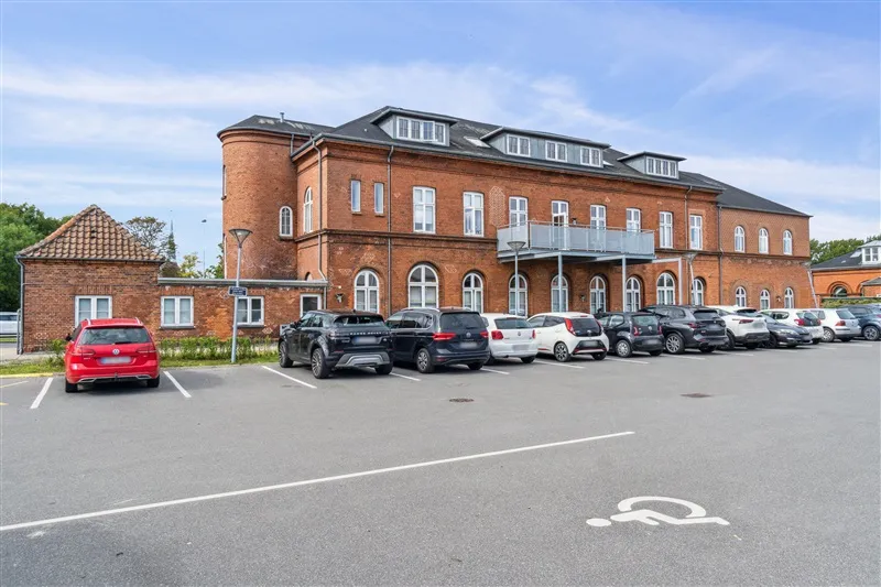 Diverse biler parkeret foran et stort bygningskompleks med mange vinduer og en indkørsel i baggrunden