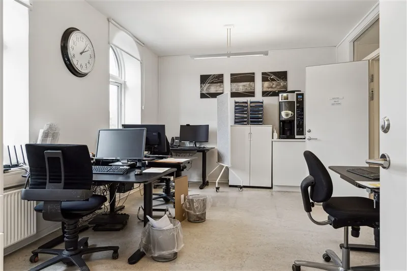 Kontor med computere, skriveborde og stolpe. Et kaffemaskine og en kalender på væggen.