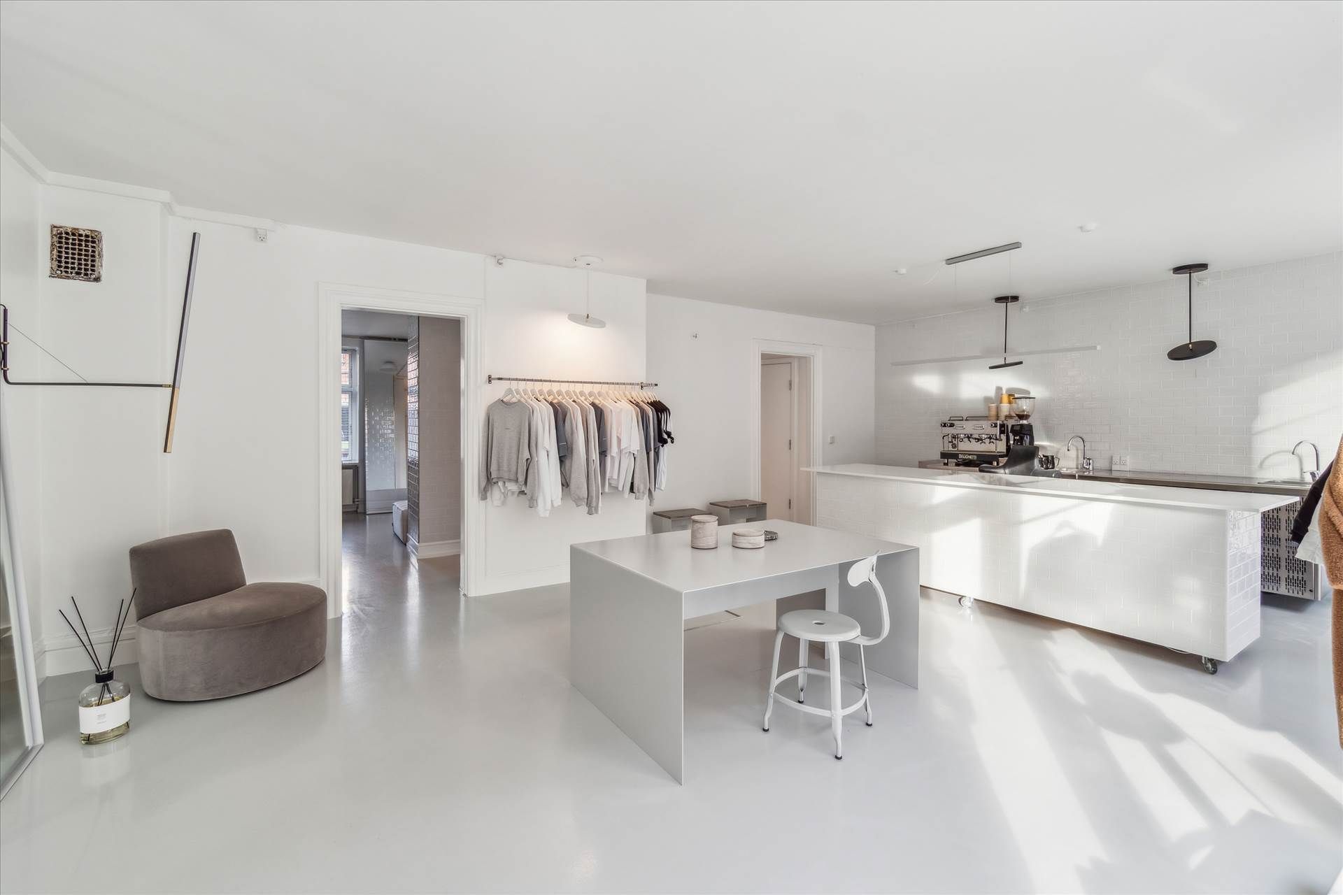 Rummelig køkken stue med hvidt loft og gulv, hængende tøj, hvidt bord med høj stol, mørkt køkken med maskine og åbne