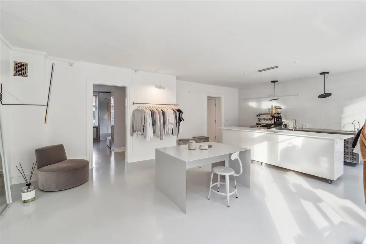 Rummelig køkken stue med hvidt loft og gulv, hængende tøj, hvidt bord med høj stol, mørkt køkken med maskine og åbne