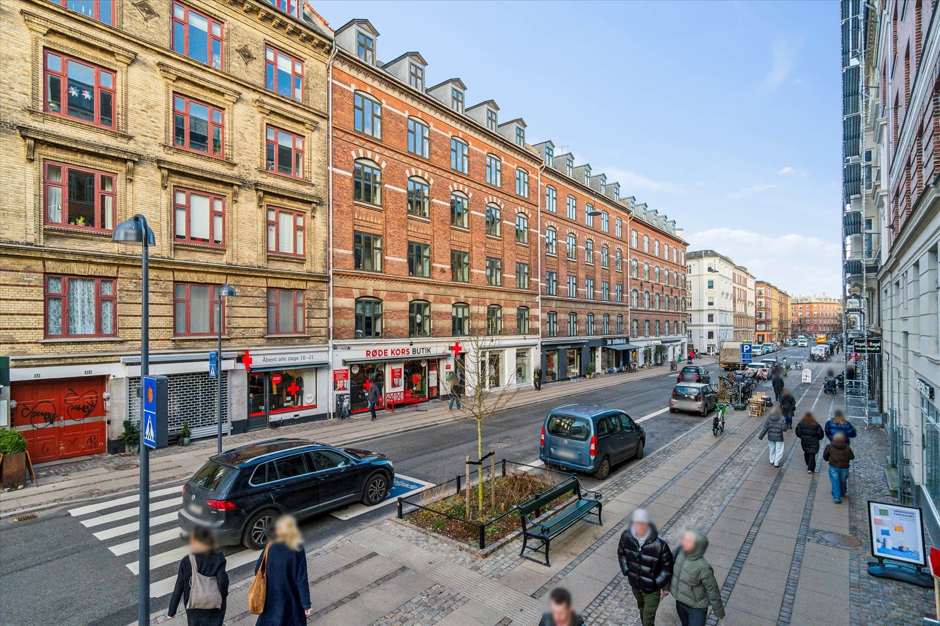 Mennesker går på gågaden med flere biler og skråbånd, facaden er røde mursten, der er et rødt skilt og en grøn plante langs